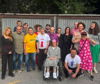 Gruppenfoto der Theatergruppe MiMe vor Garagentoren. Rund fünfzehn Personen stehen lachend beisammen, in der Mitte sitzt eine Frau im Rollstuhl, vorne kniet ein Mann.